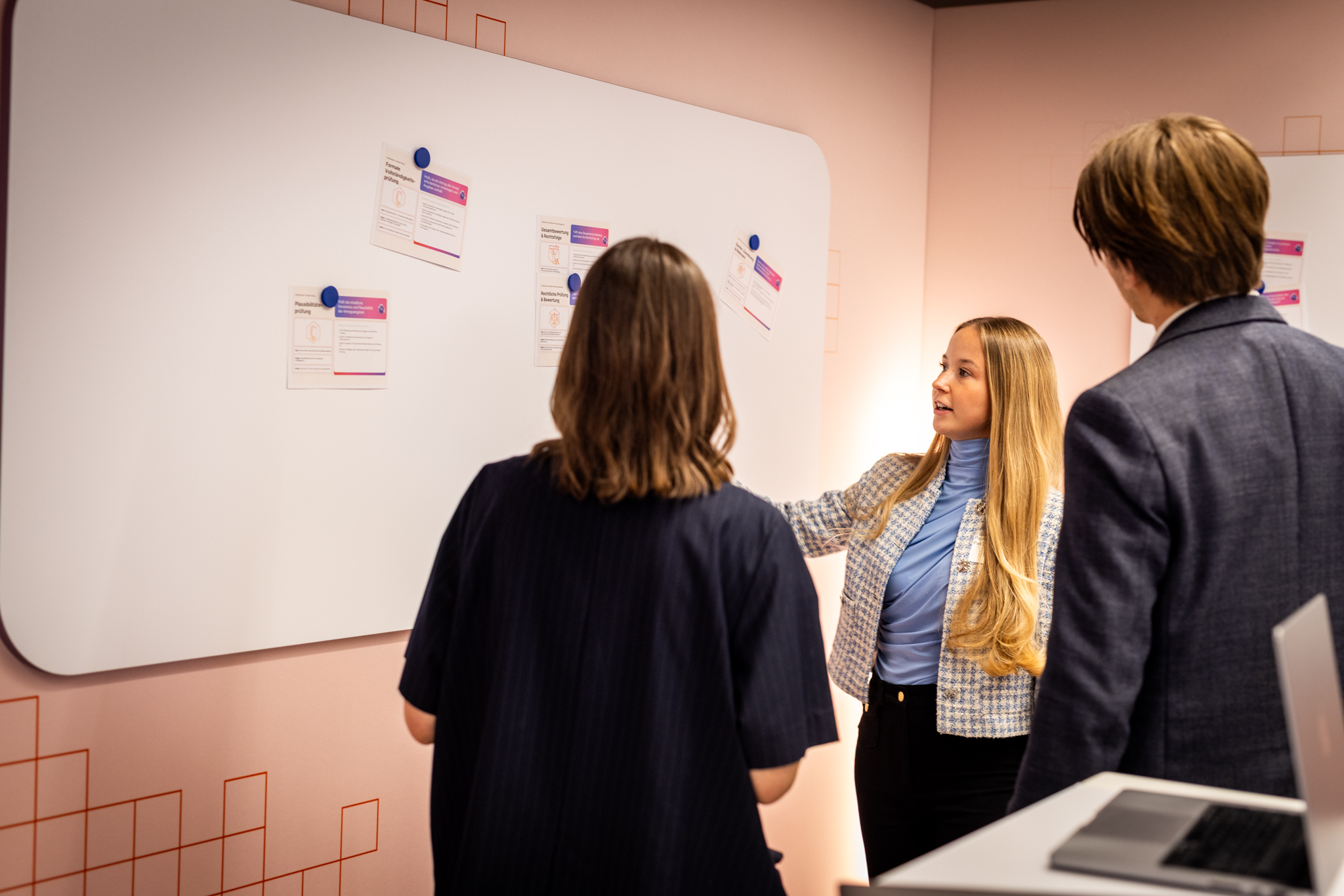 Eine Person zeigt am Whiteboard konkrete KI-Anwendungsfaelle fuer Verwaltungsprozesse, waehrend zwei weitere Teilnehmende zusehen